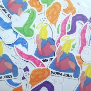 May include: A collection of colourful worm-shaped stickers with the text "Worm Jesus" on several of them. The stickers feature a variety of colours, including green, pink, purple, orange, and yellow. Some of the worms have halos and are depicted with closed eyes, suggesting they are sleeping.