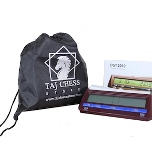 May include: A brown chess clock with a blue display and a black carrying case. The clock has two displays, one for each player, and a button for starting and stopping the clock. The carrying case has a white logo that says "Taj Chess Store" and a website address. The clock is labelled "DGT 2010".