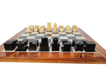 May include: A wooden chessboard with black and white squares. The board is set up with black and beige chess pieces. The pieces are in the shape of geometric blocks. The board is on a wooden table.