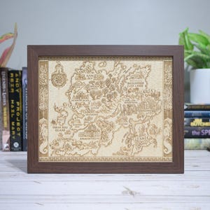 May include: A framed, detailed map with a brown wooden frame. The map is beige and features intricate illustrations of a fantasy world, including text such as "Gilaria" and "Taran Ross Island". The frame is square and sits on a white surface.