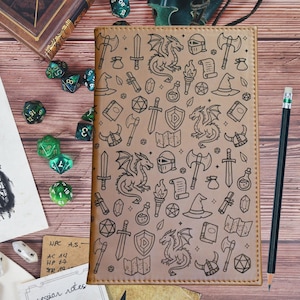 May include: A tan rectangular notebook cover featuring a repeating black line art pattern of fantasy elements including dragons, swords, axes, helmets, shields, potions, scrolls, and dice. The cover has stitched edges and a smooth texture.