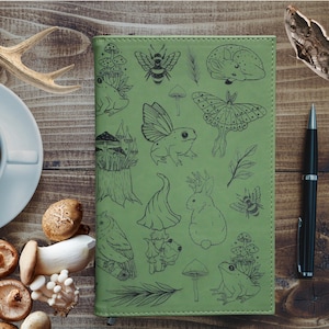 May include: A sage green notebook featuring black line art illustrations of frogs, mushrooms, a rabbit, a moth, a bee, and an owl. The cover has a smooth texture with visible stitching along the edges, showcasing a detailed nature-themed pattern.
