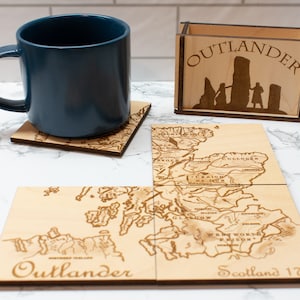 May include: A teal mug sits on a wooden coaster featuring a map design. A wooden box with the word "OUTLANDER" and a silhouette design is also present. Additional wooden coasters with a map of Scotland are also visible.