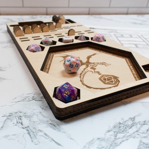 May include: A wooden dice tray with a dragon design and multiple compartments for dice. The tray is made of light wood and has a dark brown stain. There are several dice in the tray, including a pink and blue d20, a purple and blue d20, and a blue and white d6.