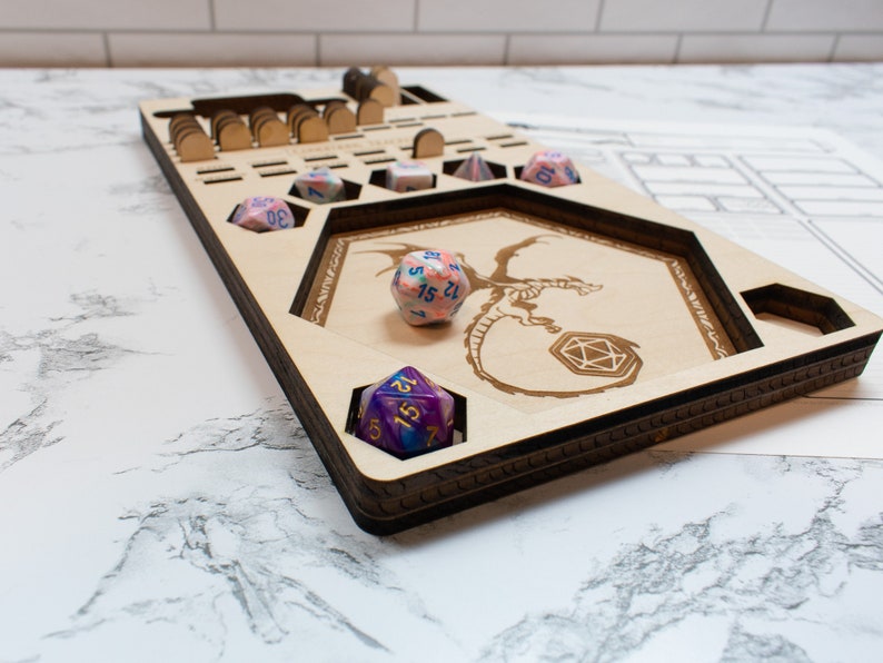May include: A wooden dice tray with a dragon design and multiple compartments for dice. The tray is made of light wood and has a dark brown stain. There are several dice in the tray, including a pink and blue d20, a purple and blue d20, and a blue and white d6.