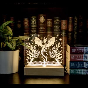 May include: A decorative LED lamp featuring a dragon design, set on a wooden base. The dragon is silhouetted against a book and floral elements. The lamp is placed on a shelf with books and a potted plant.
