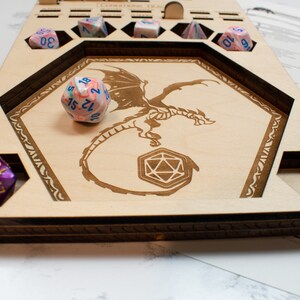 May include: Wooden dice tray with a dragon design and compartments for dice. The tray has a purple, blue, and white dice in one compartment and a pink, blue, and white dice in another compartment.