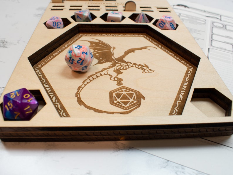May include: Wooden dice tray with a dragon design and compartments for dice. The tray has a purple, blue, and white dice in one compartment and a pink, blue, and white dice in another compartment.