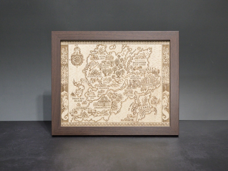 May include: A framed, detailed map with a dark brown wooden frame. The map is light brown and features intricate illustrations of a fantasy world, including landmasses, cities, and text. A compass rose is visible in the upper left corner. The frame is rectangular.