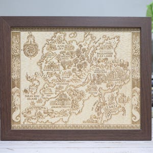 May include: A framed map with a detailed, engraved design. The map features a fictional land with names like 'Yvella' and 'Gilaria', surrounded by a dark brown wooden frame. The map is beige with brown details.