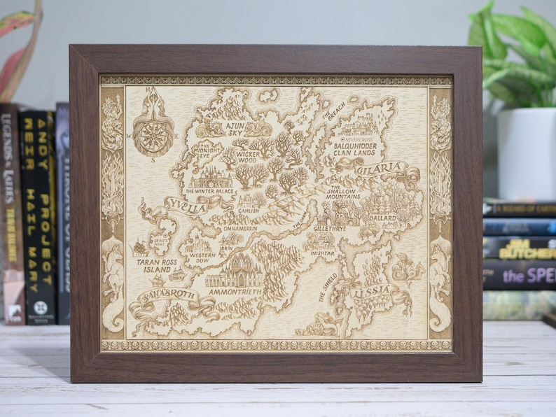 May include: A framed map with a detailed, engraved design. The map features a fictional land with names like 'Yvella' and 'Gilaria', surrounded by a dark brown wooden frame. The map is beige with brown details.