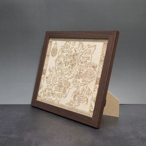 May include: A framed, detailed map with a dark brown wooden frame. The map is light brown and features intricate carvings of geographical features and text. The frame is angled, supported by a light brown stand, set against a gray background.
