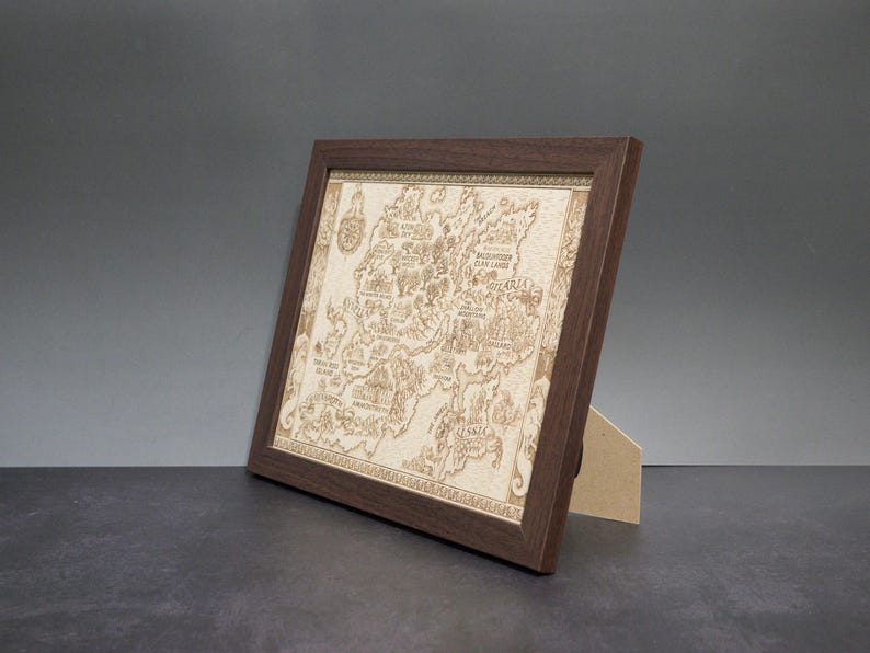 May include: A framed, detailed map with a dark brown wooden frame. The map is light brown and features intricate carvings of geographical features and text. The frame is angled, supported by a light brown stand, set against a gray background.