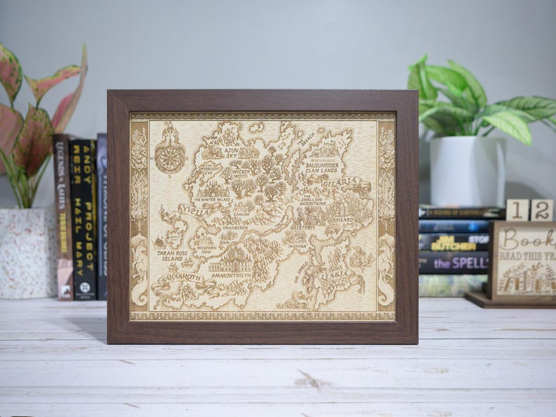 May include: A framed, detailed map with a brown wooden frame. The map is beige and features intricate illustrations of a fantasy world, including text such as "Gilaria" and "Taran Ross Island". The frame is square and sits on a white surface.