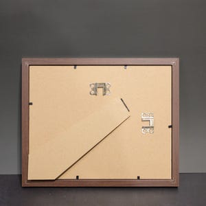 May include: Back of a brown wooden picture frame with a cardboard backing. The frame has metal clips and a hinged easel stand. The background is a dark gray color.
