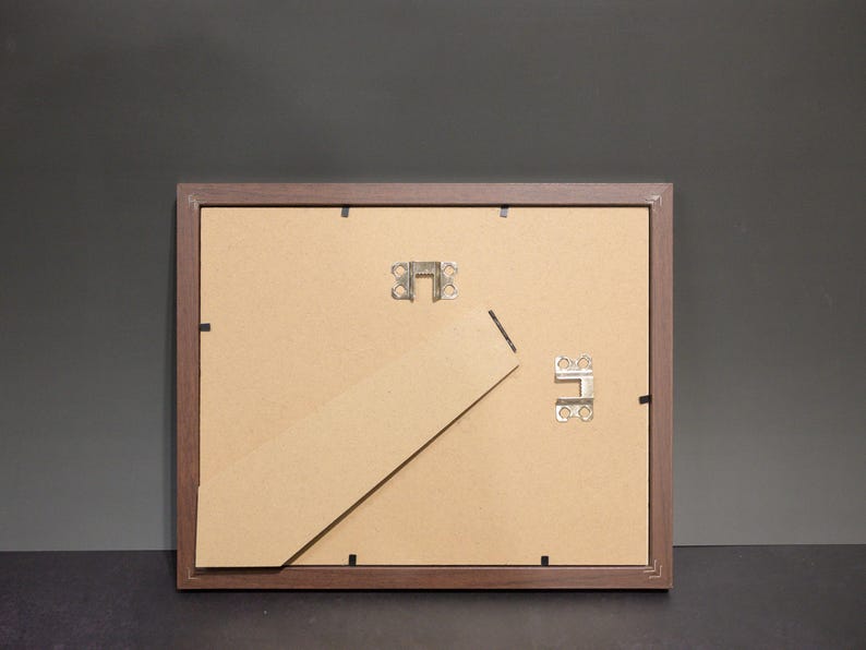 May include: Back of a brown wooden picture frame with a cardboard backing. The frame has metal clips and a hinged easel stand. The background is a dark gray color.