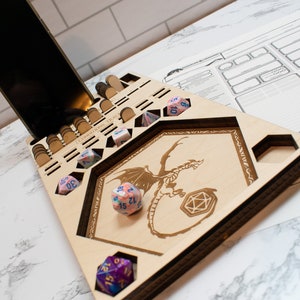 May include: A wooden dice tray with a dragon design and compartments for dice. The tray has a phone stand and a separate area for dice rolling. There are several colorful dice on the tray.