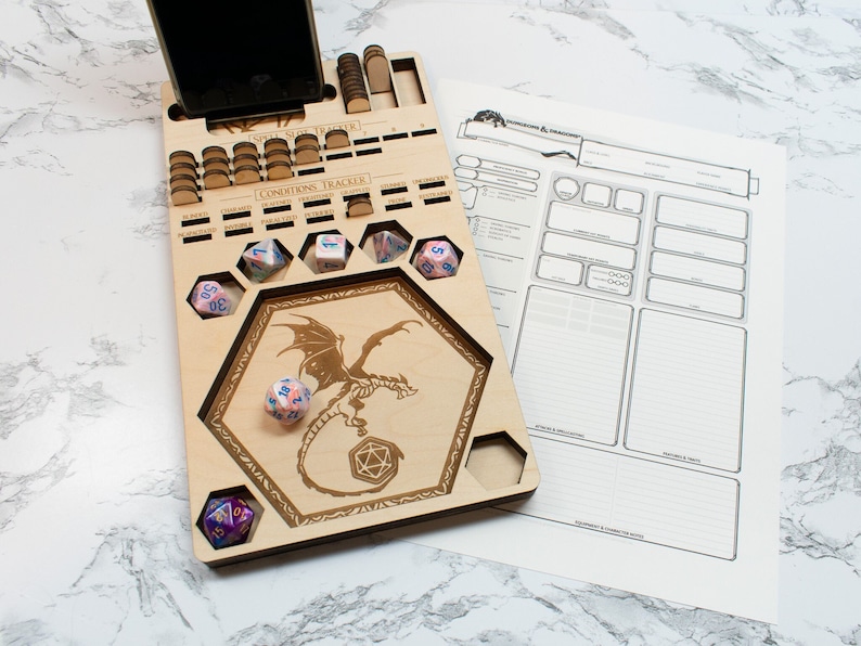 May include: A wooden game board with a dragon design and a phone slot. The board has a hexagonal area for dice and a section for tracking conditions. There are several dice on the board, including a pink and blue d20.
