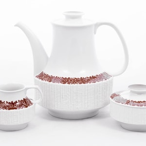 Puede incluir: Un juego de té de cerámica blanca con un diseño floral marrón. El juego incluye una tetera, una jarra de crema y un azucarero.