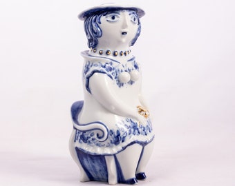 May include: A white ceramic figurine of a woman wearing a blue and white dress and a hat. She is sitting on a blue and white chair. The figurine is decorated with gold accents.