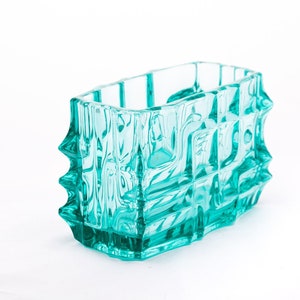 Puede incluir: Un jarrón de vidrio azul turquesa con un diseño geométrico. El jarrón tiene una forma rectangular con una superficie texturizada.