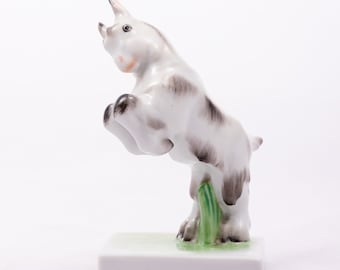 May include: A white porcelain figurine of a goat jumping up with its front legs in the air. The goat has black and gray markings and is standing on a green base.