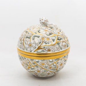 Op de afbeelding: Een decoratieve witte porseleinen container met een deksel. De bolvormige container heeft een bloemen filigraan ontwerp en gouden accenten. Het deksel heeft een kleine, decoratieve handgreep. De container wordt waarschijnlijk gebruikt voor het opbergen van kleine items of als decoratief stuk.