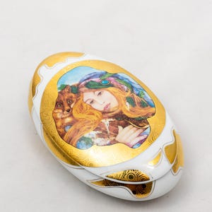 May include: An oval porcelain trinket box featuring a white base with ornate gold leaf detailing. The lid displays a colorful illustration of a person with long red hair and a fox, accented by a decorative gold border with intricate patterns.