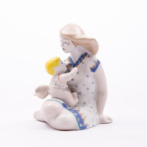 Peut inclure: Figurine en porcelaine repr&eacute;sentant une m&egrave;re et son enfant. La m&egrave;re est assise, v&ecirc;tue d'une robe blanche &agrave; pois bleus et garnitures. L'enfant, aux cheveux jaunes, tend les bras vers la m&egrave;re. La figurine a une palette de couleurs douces et pastel.