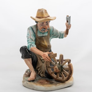 Peut inclure: Figurine en céramique représentant un artisan affûtant une hache. La figurine porte un chapeau et un tablier, et est assise avec une roue en bois. La palette de couleurs comprend des nuances de marron, de bleu et de beige.