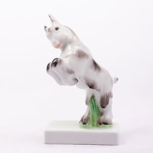 May include: A white porcelain figurine of a goat jumping up with its front legs in the air. The goat has black and gray markings and is standing on a green base.