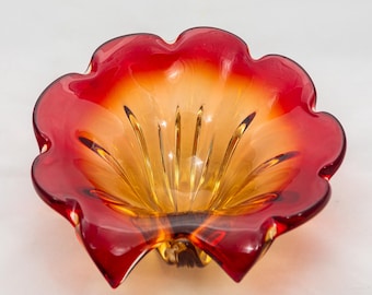 May include: A decorative glass bowl in a flower shape. The bowl features a vibrant gradient of colors, transitioning from a deep red at the edges to a warm amber in the center. The bowl has a scalloped rim and a small base.