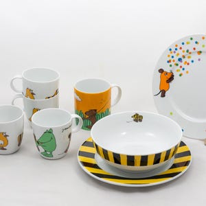 Può includere: Una collezione di stoviglie in ceramica bianca con illustrazioni di cartoni animati. Include tazze con disegni di animali, una ciotola e un piatto con strisce nere e gialle e un piatto grande con un personaggio di topo e punti colorati.