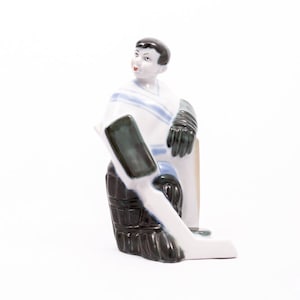 May include: A porcelain figurine of a hockey goalie in a white, blue, and black uniform, sitting with a stick in hand.