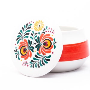 May include: A white ceramic jar with a red stripe and a floral design on the lid. The lid is open and a small black Lego block is on the table.