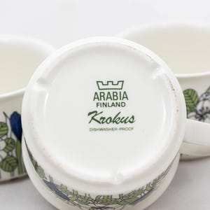 May include: Three white ceramic mugs with floral designs in blue, green, and white. The bottom of one mug displays the text "ARABIA FINLAND Krokus DISHWASHER-PROOF". The mugs have handles and are set against a white background.