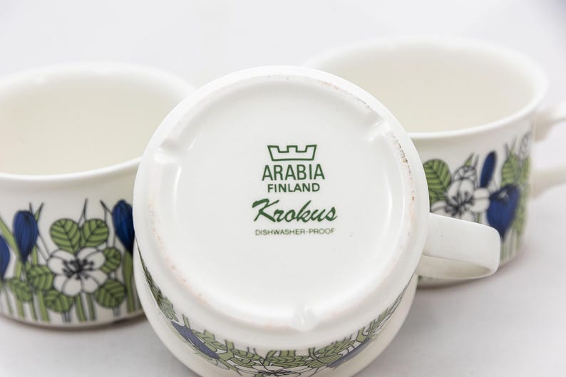 May include: Three white ceramic mugs with floral designs in blue, green, and white. The bottom of one mug displays the text "ARABIA FINLAND Krokus DISHWASHER-PROOF". The mugs have handles and are set against a white background.