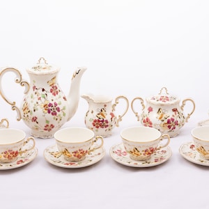 Puede incluir: Juego de té vintage sobre fondo blanco. El juego incluye una tetera, una jarra de crema, un azucarero y seis tazas y platillos. Cada pieza está decorada con motivos florales y de mariposas en rosa, amarillo y detalles dorados.