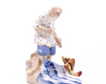 May include: A porcelain figurine depicting an elderly man in a white shirt with blue polka dots and blue shorts, looking at a golden fish. The base features blue and white waves and gold accents.