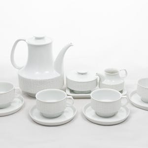 Puede incluir: Juego de té de cerámica blanca con un diseño de rayas verticales texturizadas. El juego incluye una tetera, azucarero, lechera y varias tazas de té con platillos. Las piezas están dispuestas.