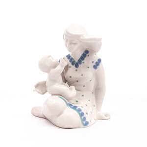 Peut inclure: Figurine en c&eacute;ramique blanche repr&eacute;sentant une m&egrave;re et son enfant. La m&egrave;re est assise, v&ecirc;tue d'une robe &agrave; pois et bordures bleus. L'enfant est tenu dans ses bras. Objet d&eacute;coratif.