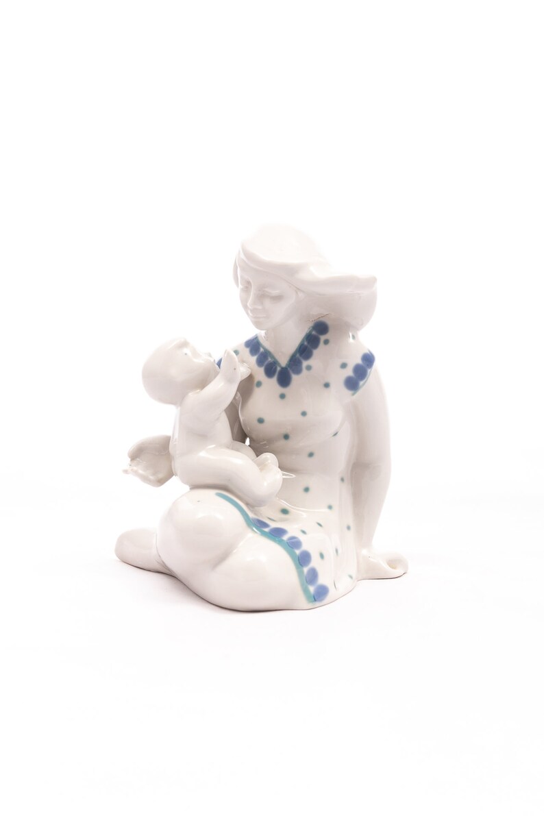 Peut inclure: Figurine en c&eacute;ramique blanche repr&eacute;sentant une m&egrave;re et son enfant. La m&egrave;re est assise, v&ecirc;tue d'une robe &agrave; pois et bordures bleus. L'enfant est tenu dans ses bras. Objet d&eacute;coratif.