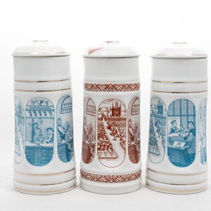 Op de afbeelding: Drie witte keramische bierpullen met deksels, elk versierd met gedetailleerde illustraties in blauw en bruin. De pullen hebben gouden randen en tonen scènes in gewelfde kaders. Een klassiek verzamelobject.