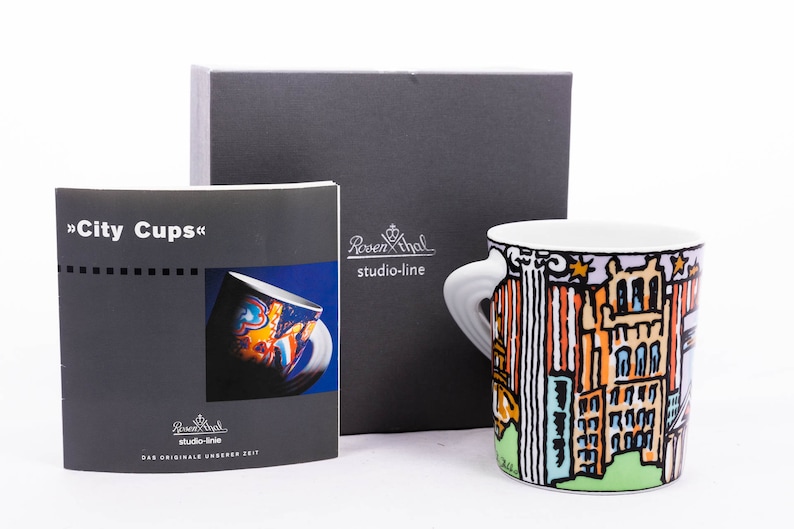May include: A white ceramic mug featuring a colourful illustration of a city skyline. The mug has two handles and is part of a set called "City Cups". The mug is packaged in a grey box with the brand name "Rosenthal studio-line" printed on it.