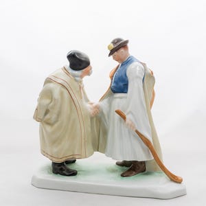 Puede incluir: Figura de porcelana que representa a dos hombres dándose la mano. Uno lleva sombrero negro y túnica crema, el otro chaleco azul, falda blanca y sostiene un bastón de madera. La base es blanca con una superficie verde y blanca.