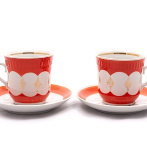 May include: Two white teacups with red and gold trim and matching saucers. The cups and saucers have a white background with a red band and gold geometric designs.