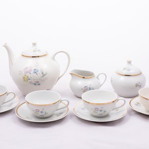 Puede incluir: Un juego de té de porcelana blanca con un diseño floral y ribete dorado. El juego incluye una tetera, una jarra de crema, un azucarero y cuatro tazas de té y platillos.
