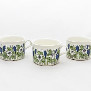 May include: Three white ceramic mugs with a floral pattern. The design features blue, green, and white flowers and foliage. Each mug has a handle and a slightly flared rim. The mugs are arranged on a white surface.