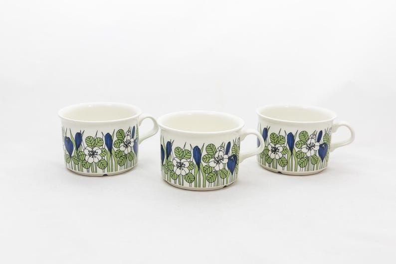 May include: Three white ceramic mugs with a floral pattern. The design features blue, green, and white flowers and foliage. Each mug has a handle and a slightly flared rim. The mugs are arranged on a white surface.