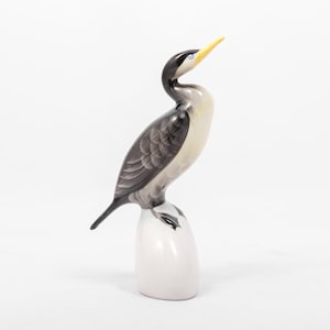 May include: A porcelain figurine of a black and white cormorant with a yellow beak standing on a white base.